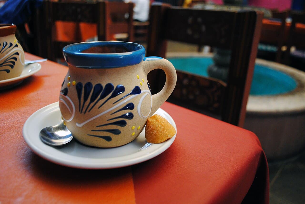 Café de olla served in a painted clay jarrito with a lump of panela on the saucer, rendered in ink and gold duotone.